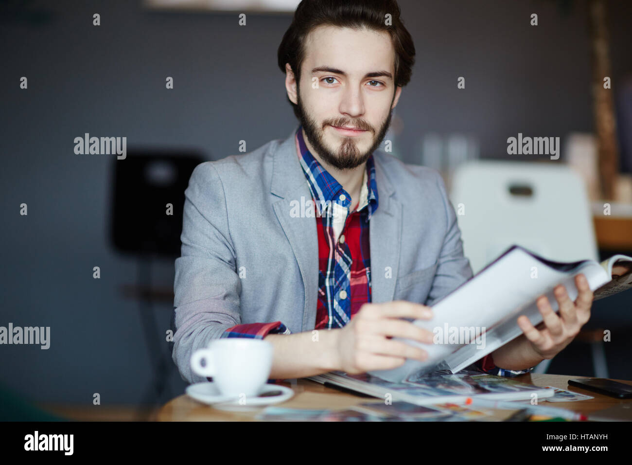Portrait of young long haired man wearing business casual clothes sitting at table looking at camera and smiling while reading magazines Stock Photo