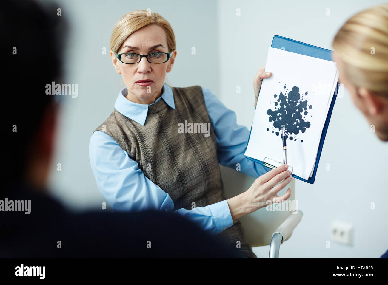 Portrait of mature female psychologist wearing glasses showing Rorschach test card to her patient in therapy session Stock Photo