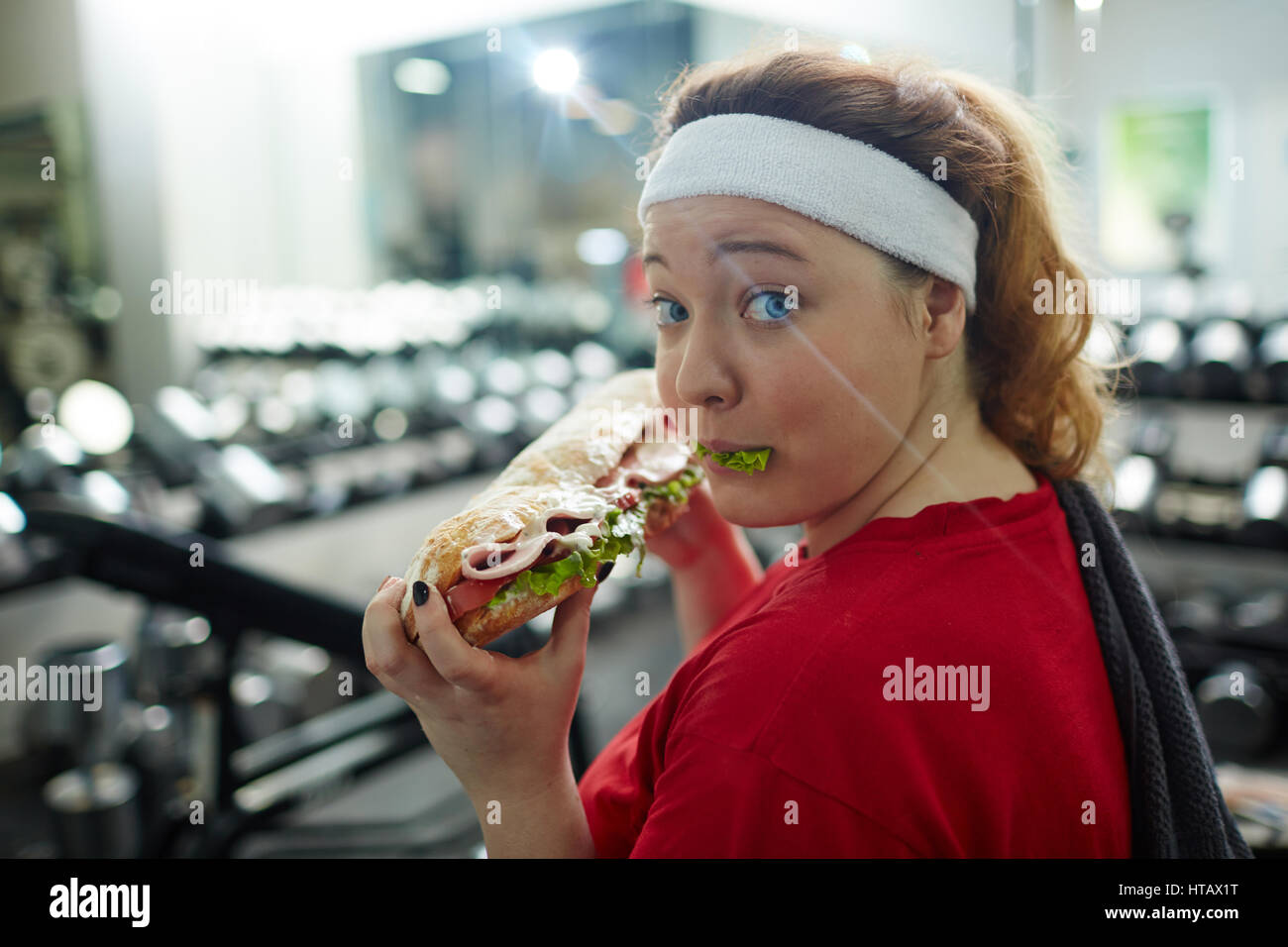 Portrait of overweight woman looking at camera, while eating  huge fattening sandwich while working out in gym, struggle of weight loss when dieting Stock Photo