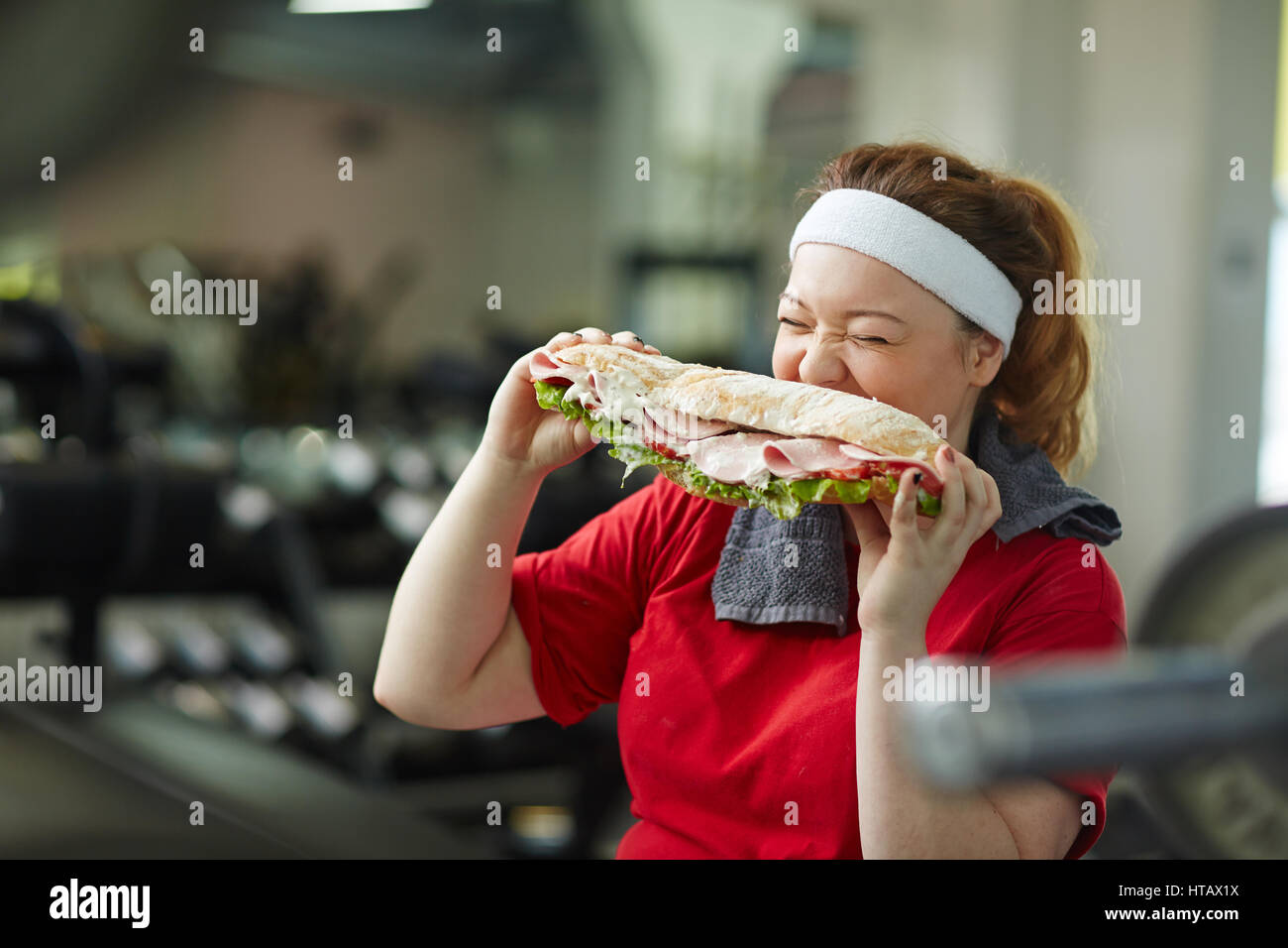 Portrait of young overweight woman eating big fattening sandwich taking a break from work out in gym, concept of food obsession Stock Photo