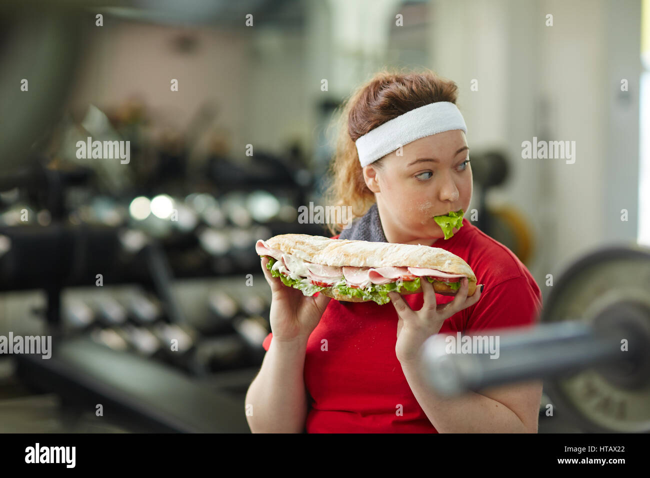 Portrait of young overweight woman eating big greasy fattening sandwich at work out in gym, concept of food obsession Stock Photo