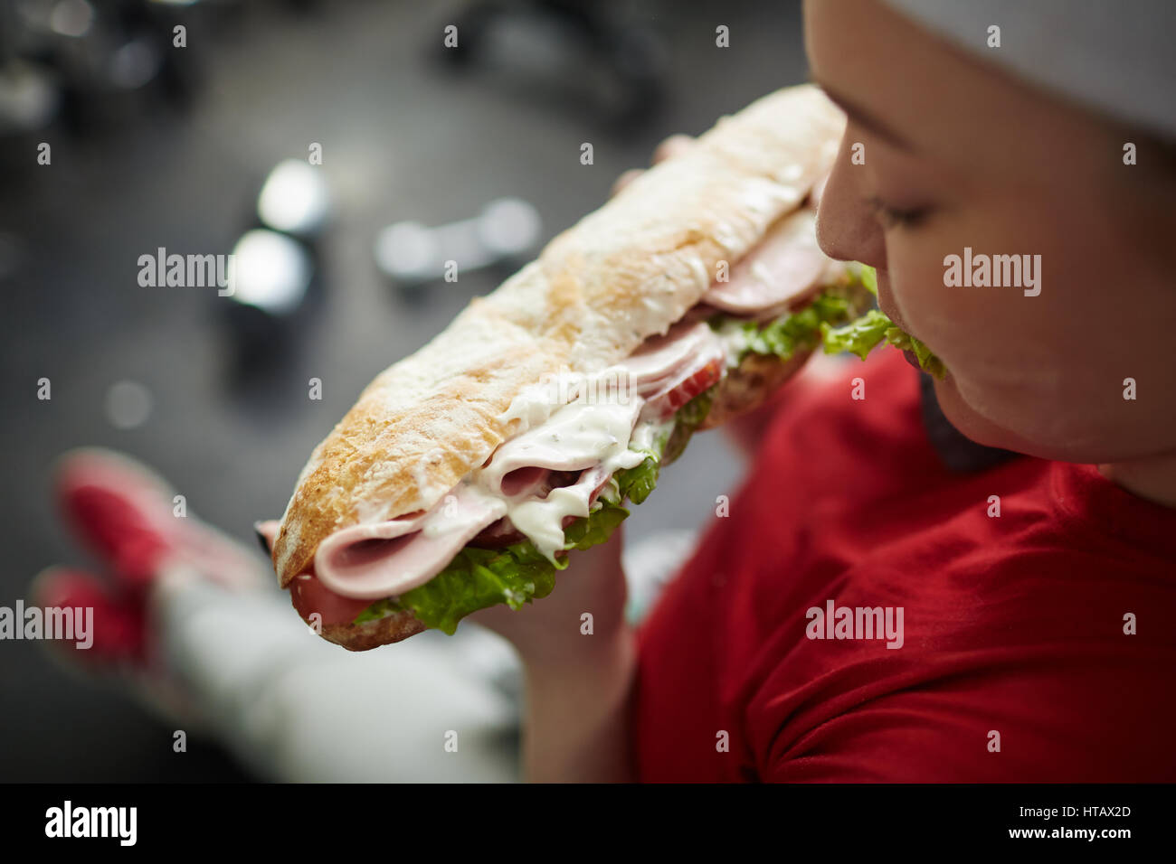 High angle shot of young overweight woman eating big greasy fattening sandwich at work out in gym, concept of food obsession Stock Photo