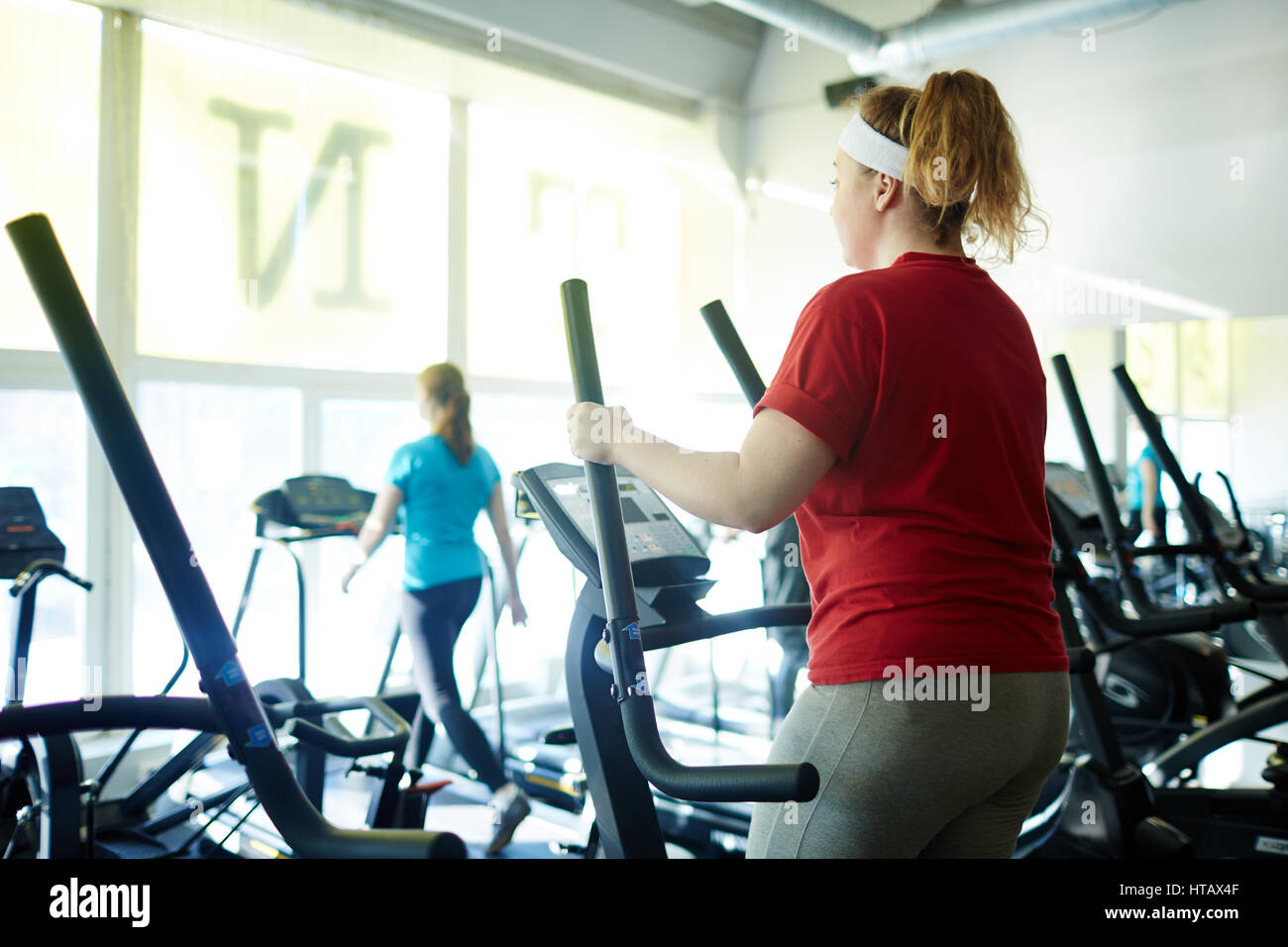 Back view  portrait of young obese woman working out in gym: using ellipse  machine with effort to lose weight Stock Photo