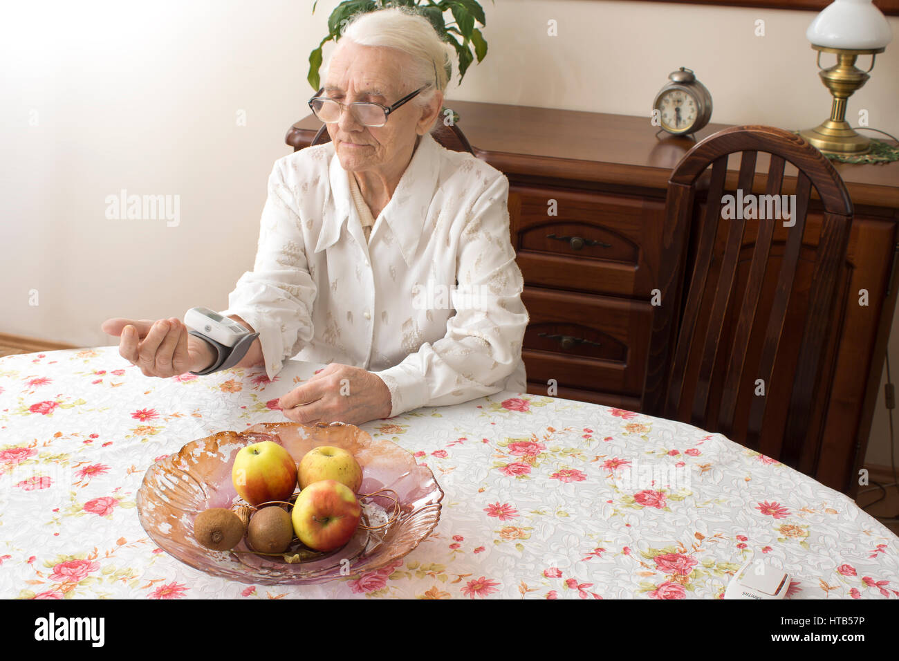 The old woman measures arterial pressure while sitting in the living room at the table. The old woman measures the pressure apparatus wrist. Stock Photo