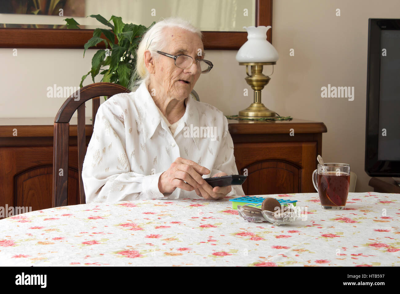Old woman at the table with remote control for the TV.  The old woman turns on the TV remote control sitting at the table.  The old woman in glasses s Stock Photo