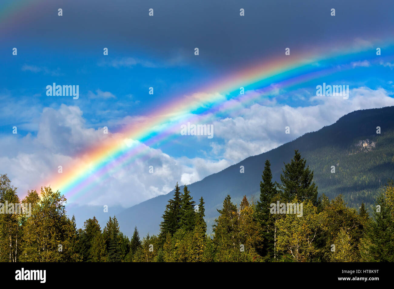 Colourful rainbow across the sky with mountain side, storm clouds and blue sky in the background; Revelstoke, British Columbia, Canada Stock Photo
