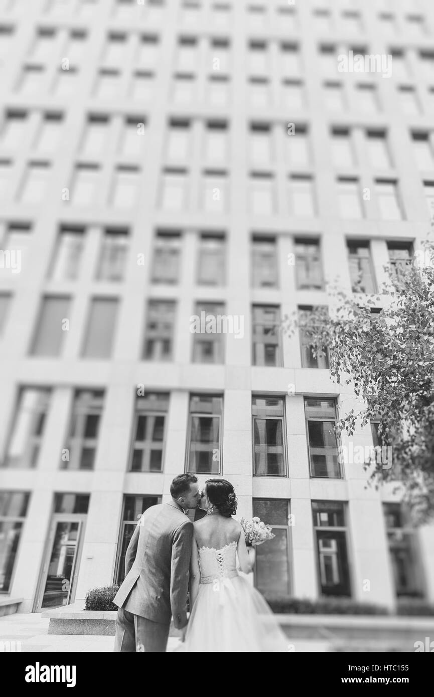 Back view young kissing and embracing couple outdoors. Bride and groom look at each other. Building in the background. Bouquet in hand. Black and whit Stock Photo
