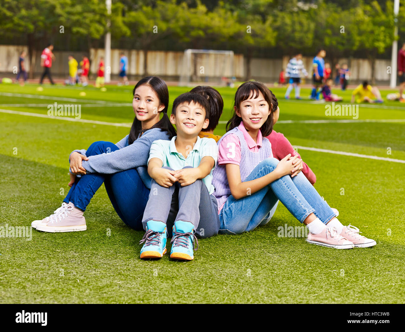 group of happy smiling elementary school boys and girls sitting on grass of playground. Stock Photo