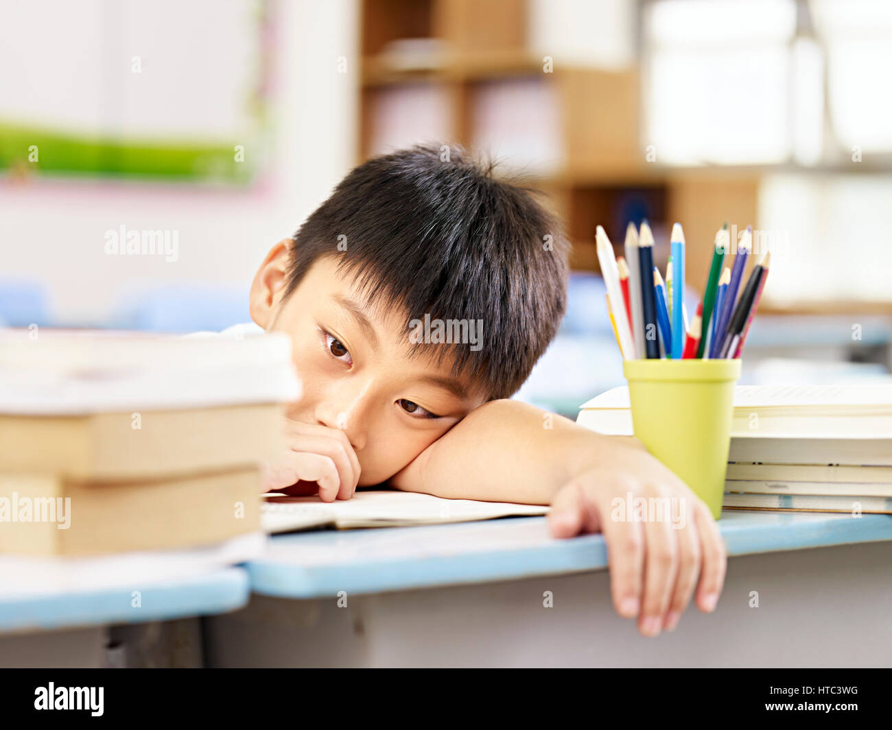 asian elementary schoolboy looking tired and exhausted resting his head on desk in classroom. Stock Photo