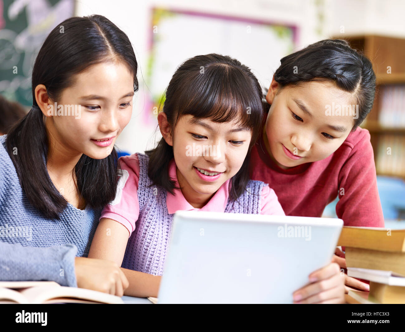 three asian elementary school girls friends looking at a tablet together during break in classroom. Stock Photo