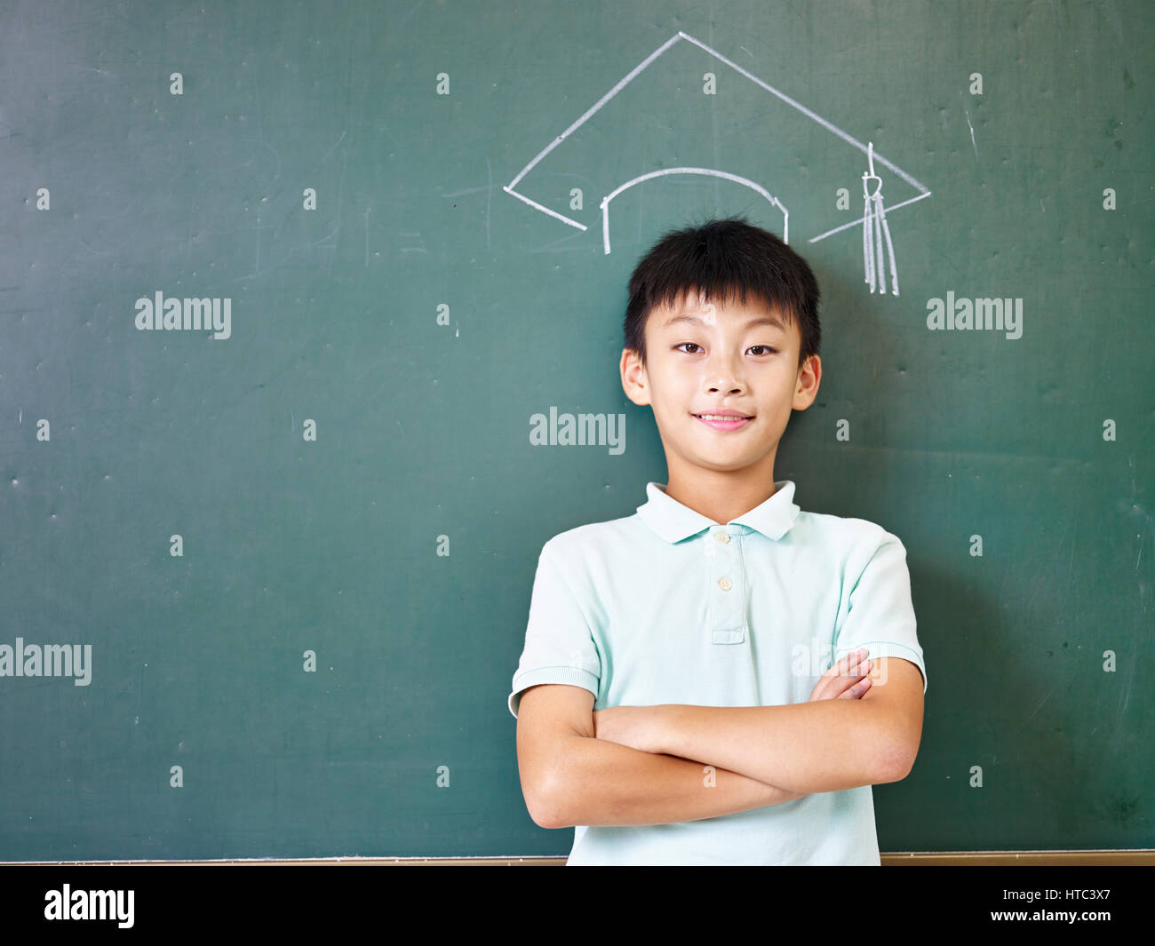 asian elementary school boy standing under a doctoral hat drawn with chalk on blackboard. Stock Photo