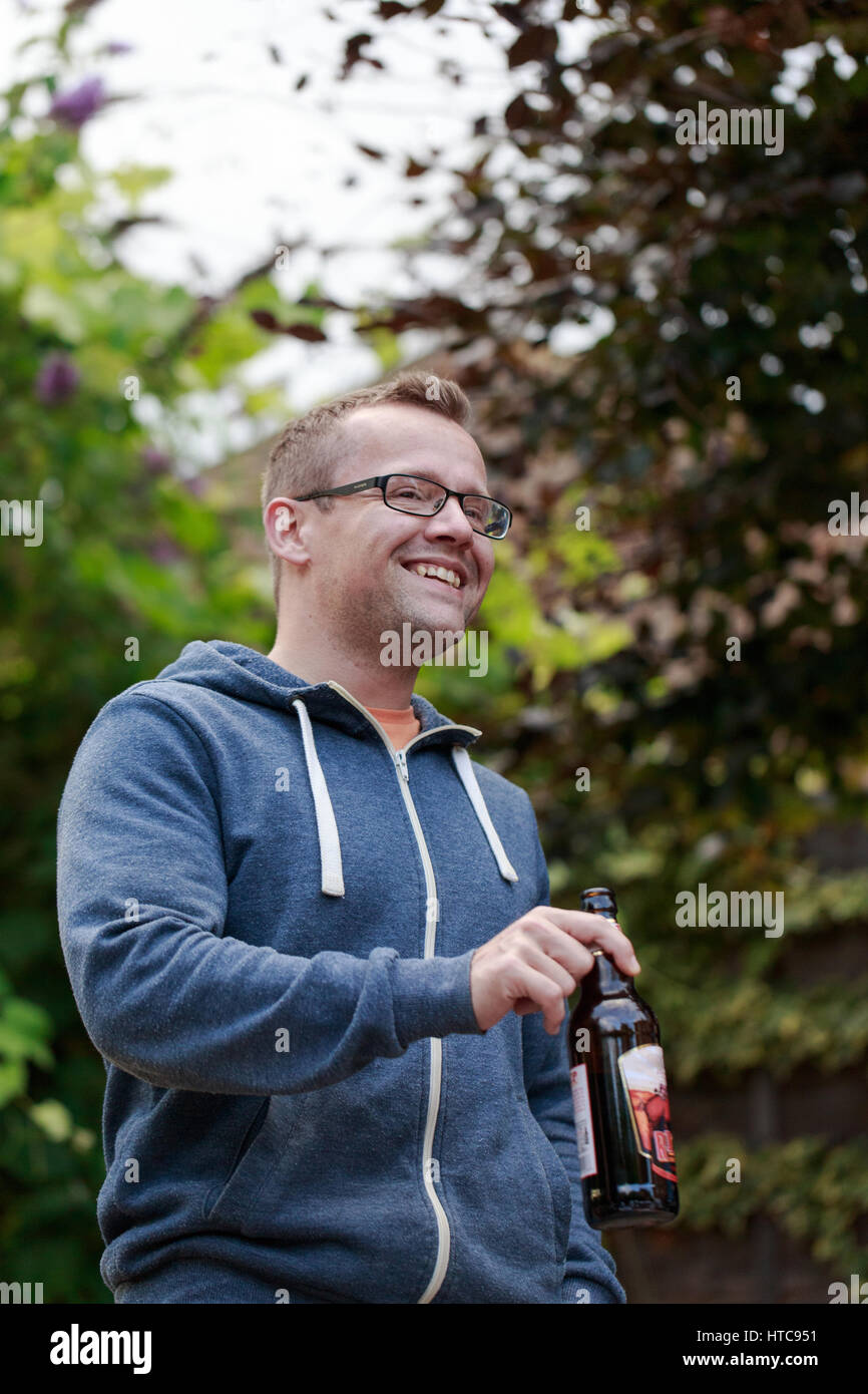Young short blond hair man wearing glasses and blue hoodie jumper drinking ale. Friends meeting after work, BBQ in garden. Vertical shot Stock Photo