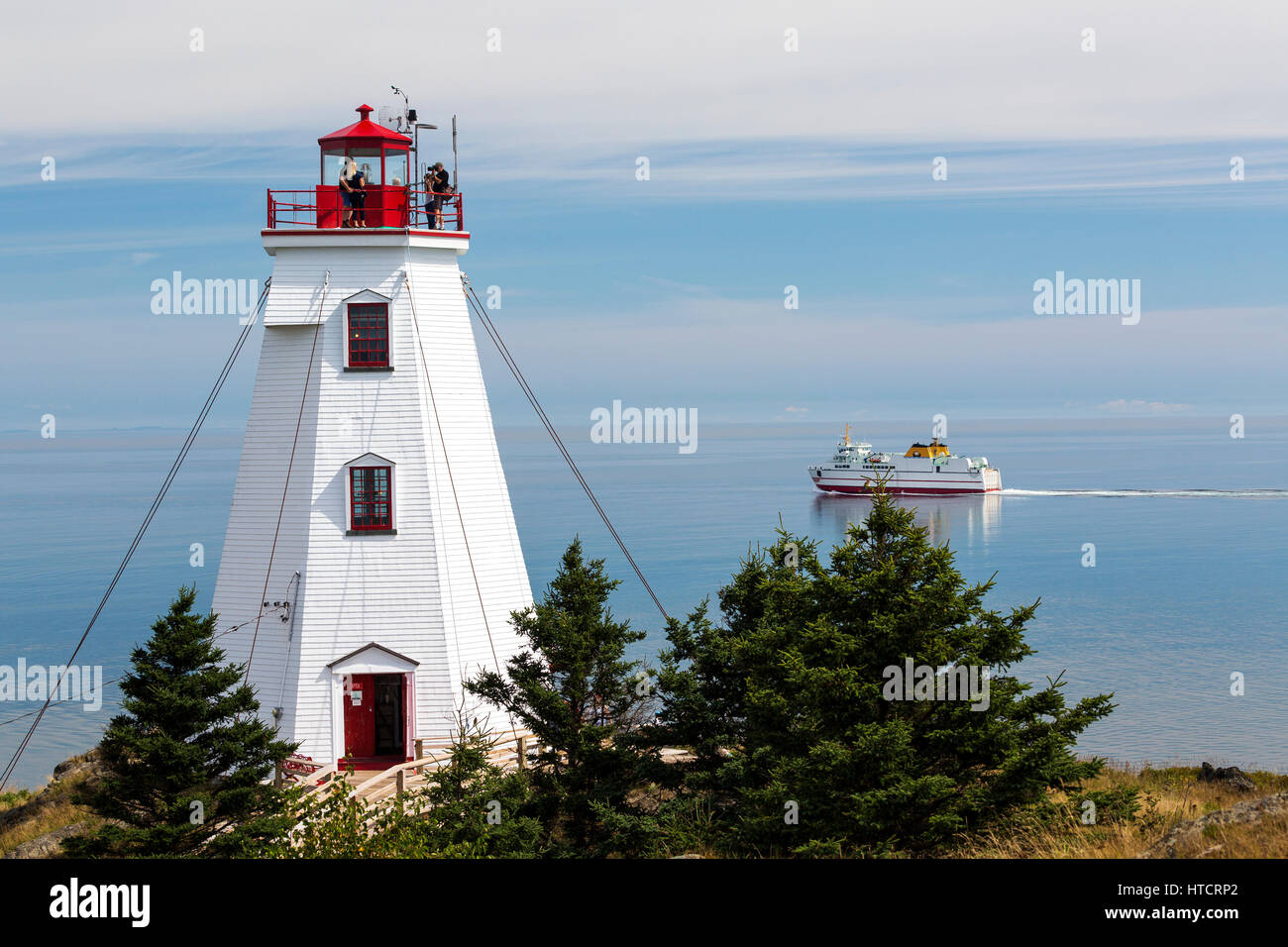 White lighthouse with people on top platform overlooking ferry crossing in the distance with hazy blue sky; North Head, New Brunswick, Canada Stock Photo