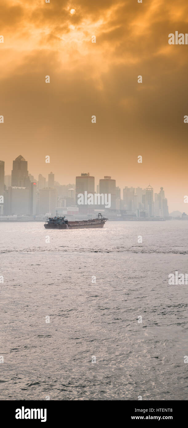 A Chinese Cargo Ship in the Victoria Harbour with the skyscrapers and the Hong Kong skyline, with the sun shining through dramatic clouds. China. Stock Photo