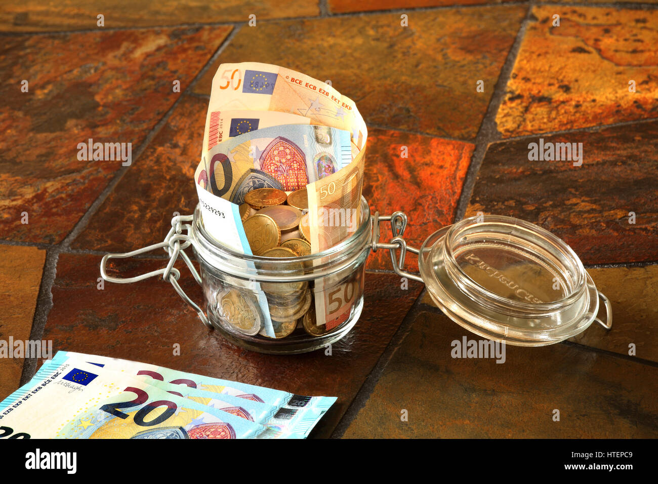 Euro banknotes and coins in a glass container with some banknotes in the left hand corner, all standing on a tiled table top Stock Photo