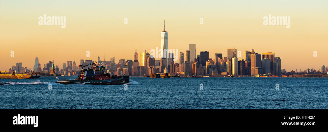 Panoramic Sunset of Lower Manhattan and New York City Harbor with Financial District skyscrapers and passing tugboat Stock Photo