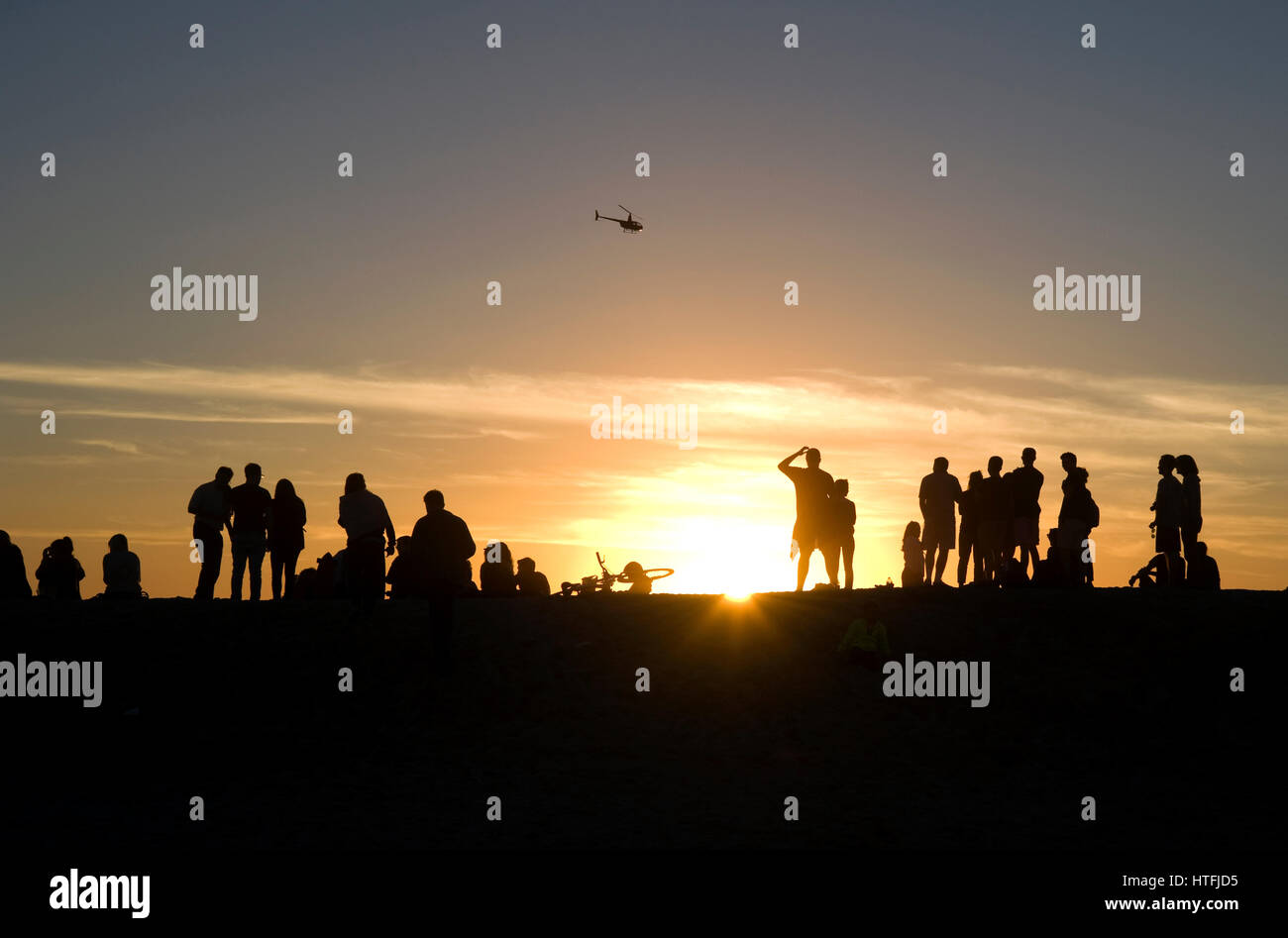 People watching the sunset at Venice Beach, California as a coast guard helicopter flies past. Stock Photo