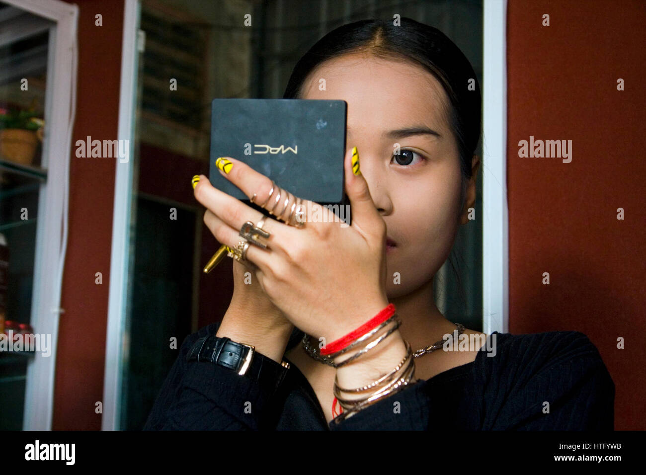 A young Asian woman is applying makeup to her face while looking in a mirror in a beauty shop in Chork Village, Cambodia. Stock Photo