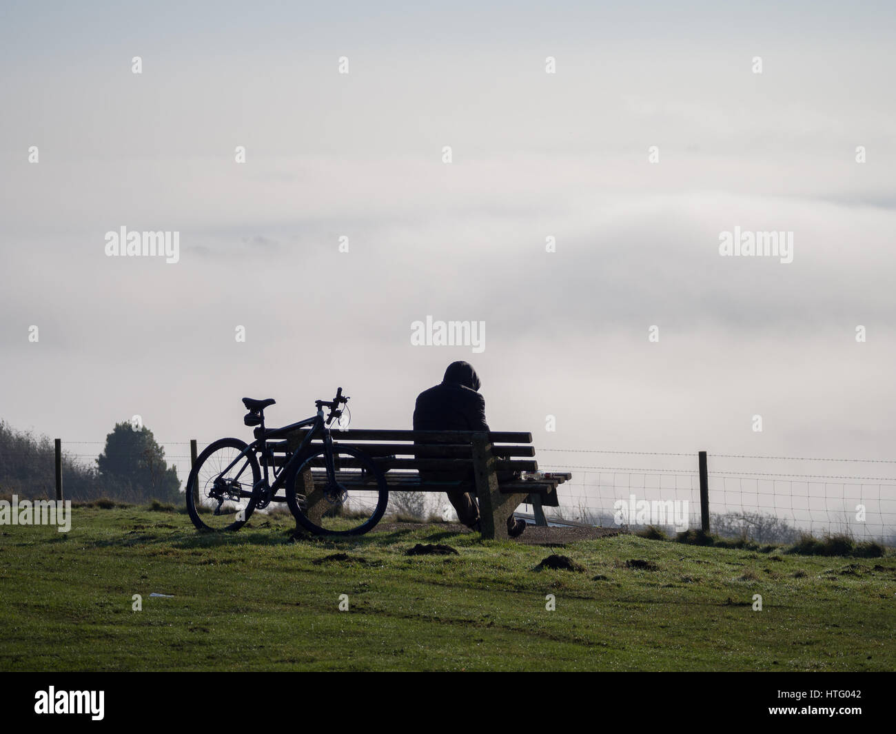 a solitary hooded cyclist silhouette sitting alone on bench on top of the Downs hill looking out down the fog mist in the valley below Stock Photo