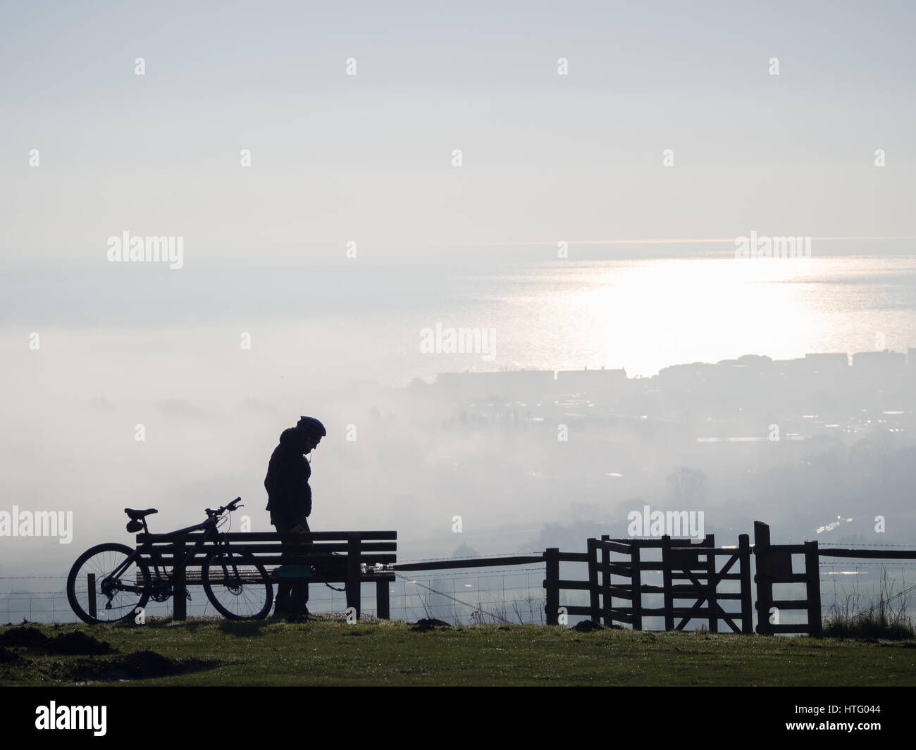 a solitary cyclist helmet silhouette profile standing alone on top of Downs hill with style looking out down the fog mist in the valley and sea below Stock Photo