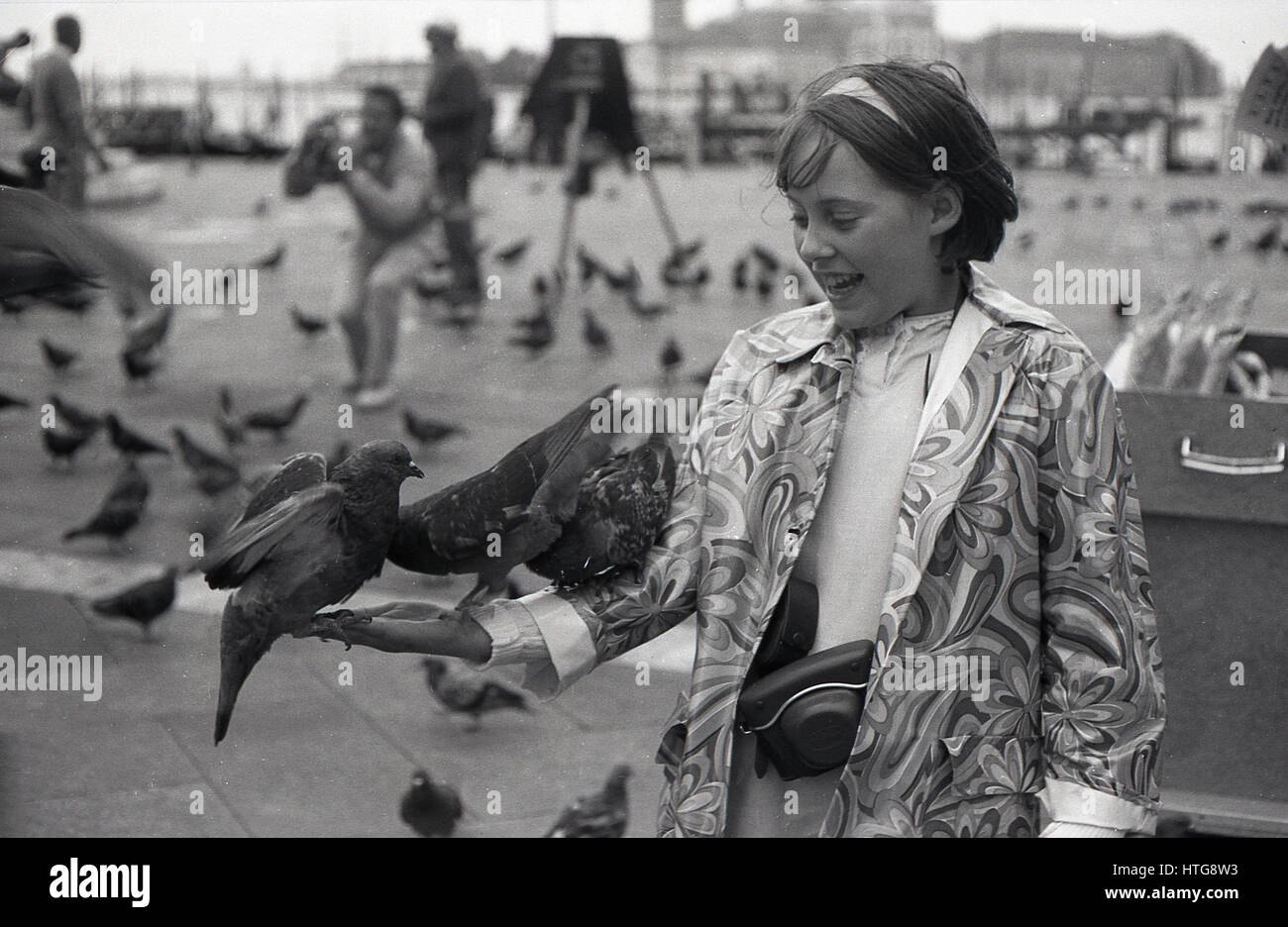 1970s, St Mark's Square. Venice, Italy, excited girl wearing a flower patterned coat standing with pigeons on her hand and arm. Stock Photo