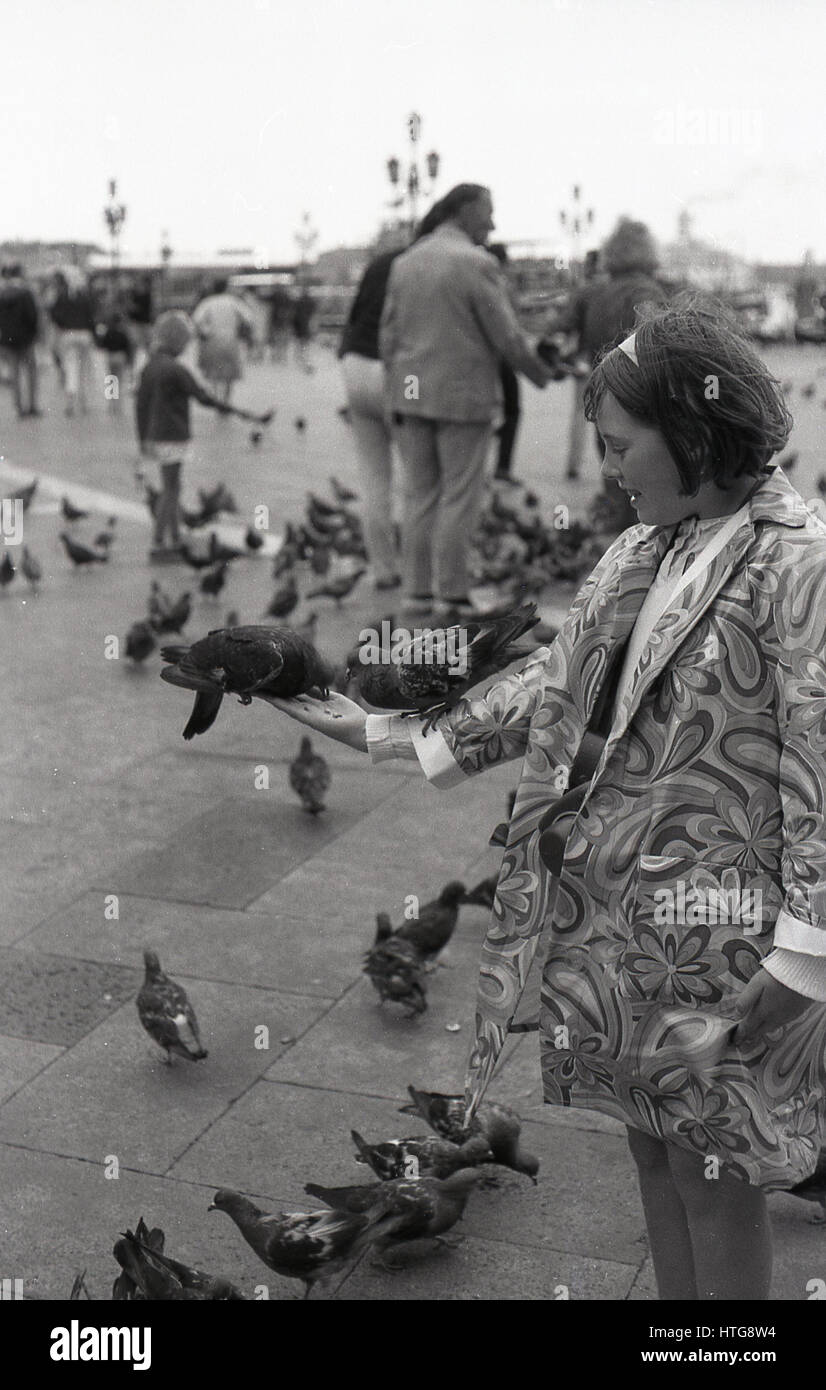 1970, historical, a girl in a flower patterned coat standing with two pigeons on her arm at St Mark's Square, Venice, Italy. A public square, it is one of Venice's prime tourist attractions. Stock Photo