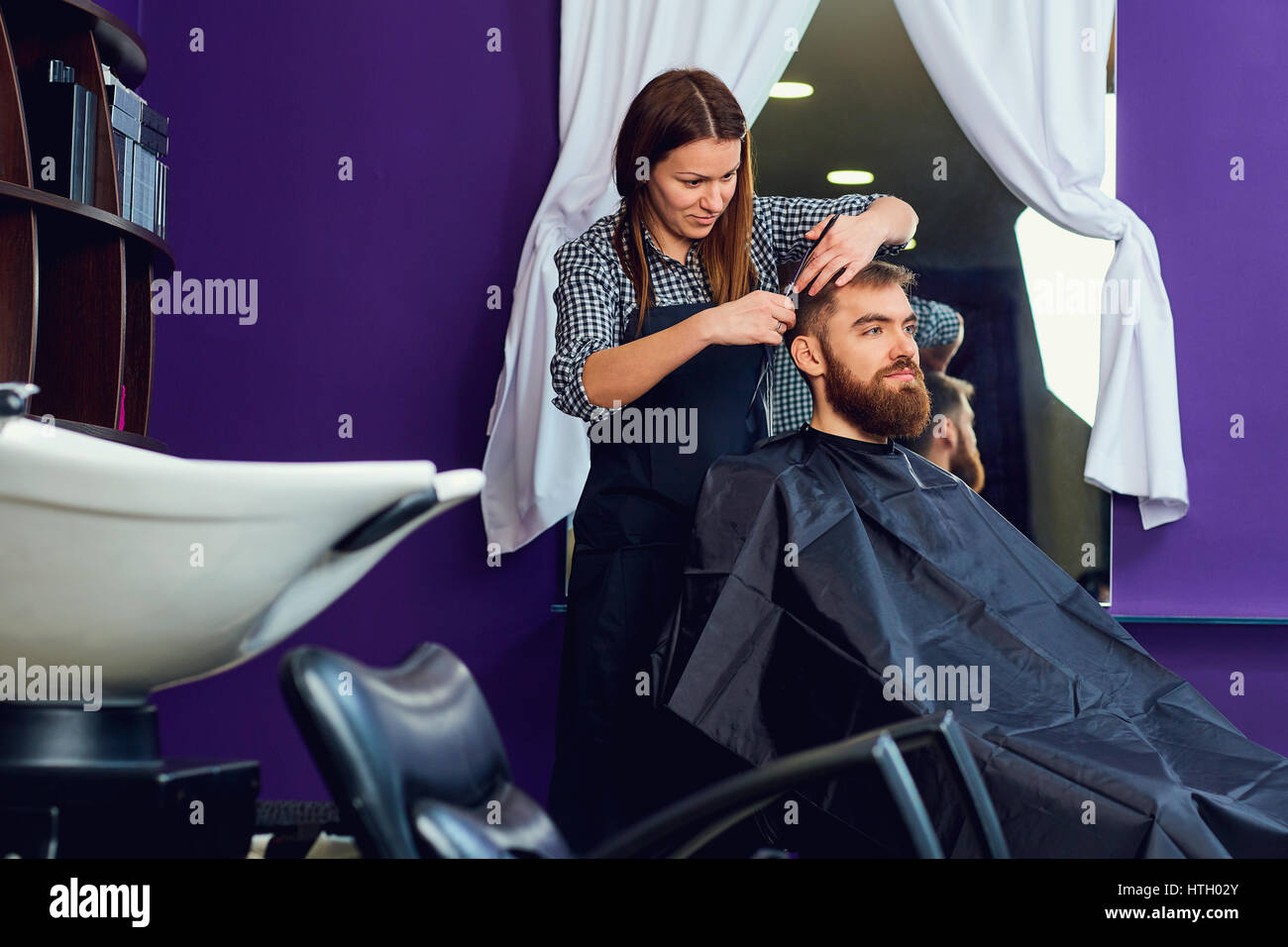 A woman hairdresser cuts  beard to  man in  beauty salon. Stock Photo