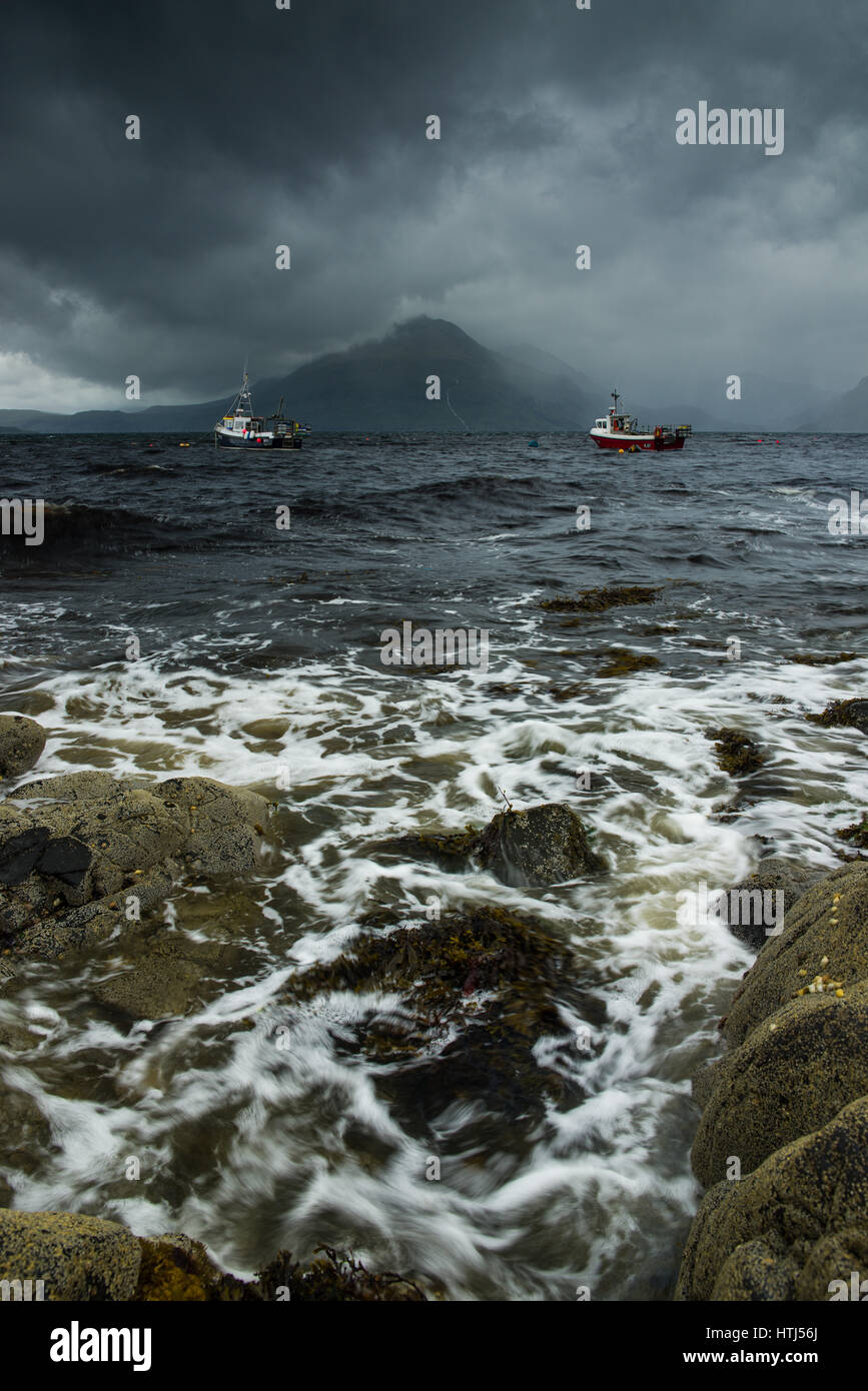 A Summer storm passes over the Cuilin Mountains and fishing boats in the sea, as seen from the rocky coast of Elgol, Isle Of Skye, Scotland Stock Photo