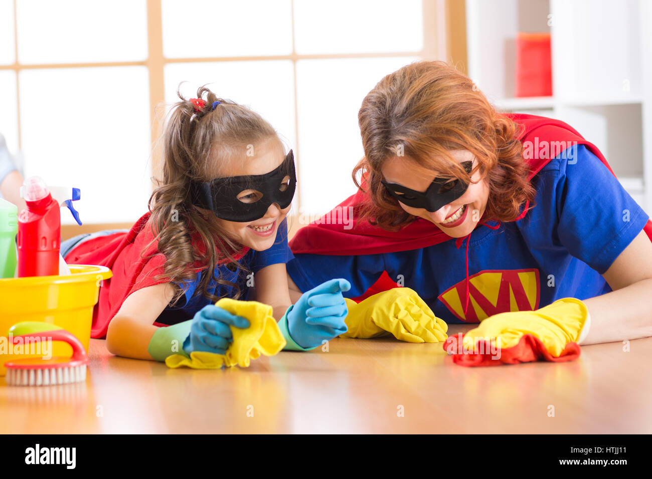 Cute woman and her kid daughter dressed like superheroes cleaning the floor and smiling Stock Photo