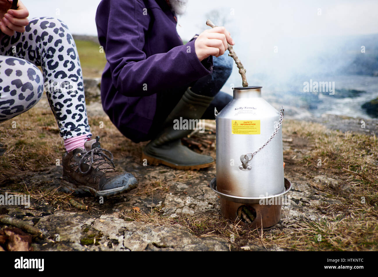 Two children outdoors around a Kelly Kettle wood burning stove kettle feeding in wood Stock Photo
