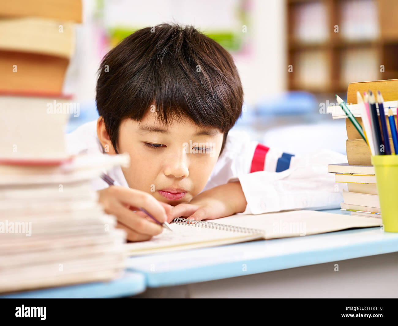 tired and bored asian elementary school boy doing homework in classroom, head resting on desk. Stock Photo