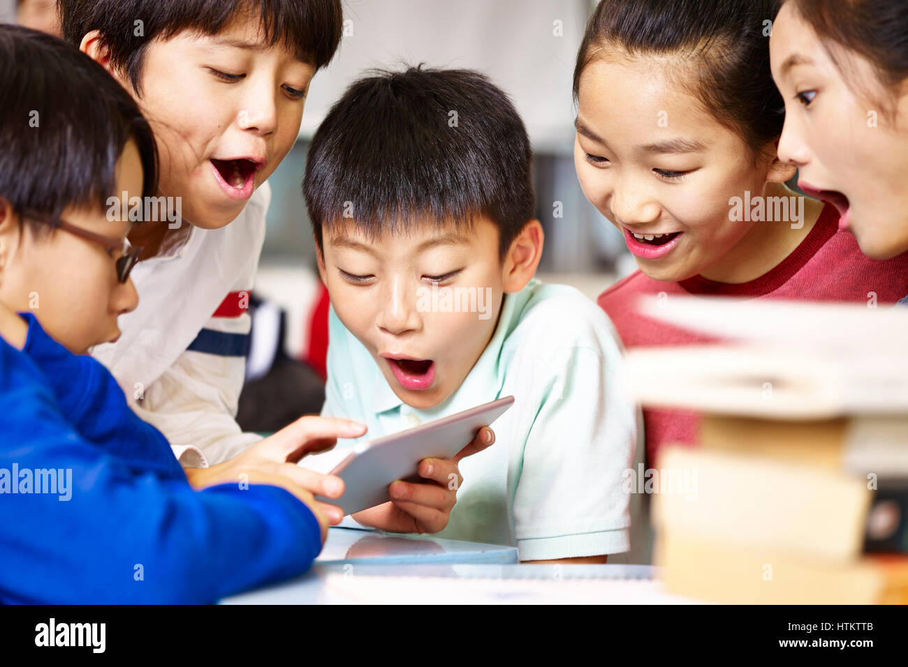 group of asian elementary school children gathering around playing game together using tablet during break. Stock Photo