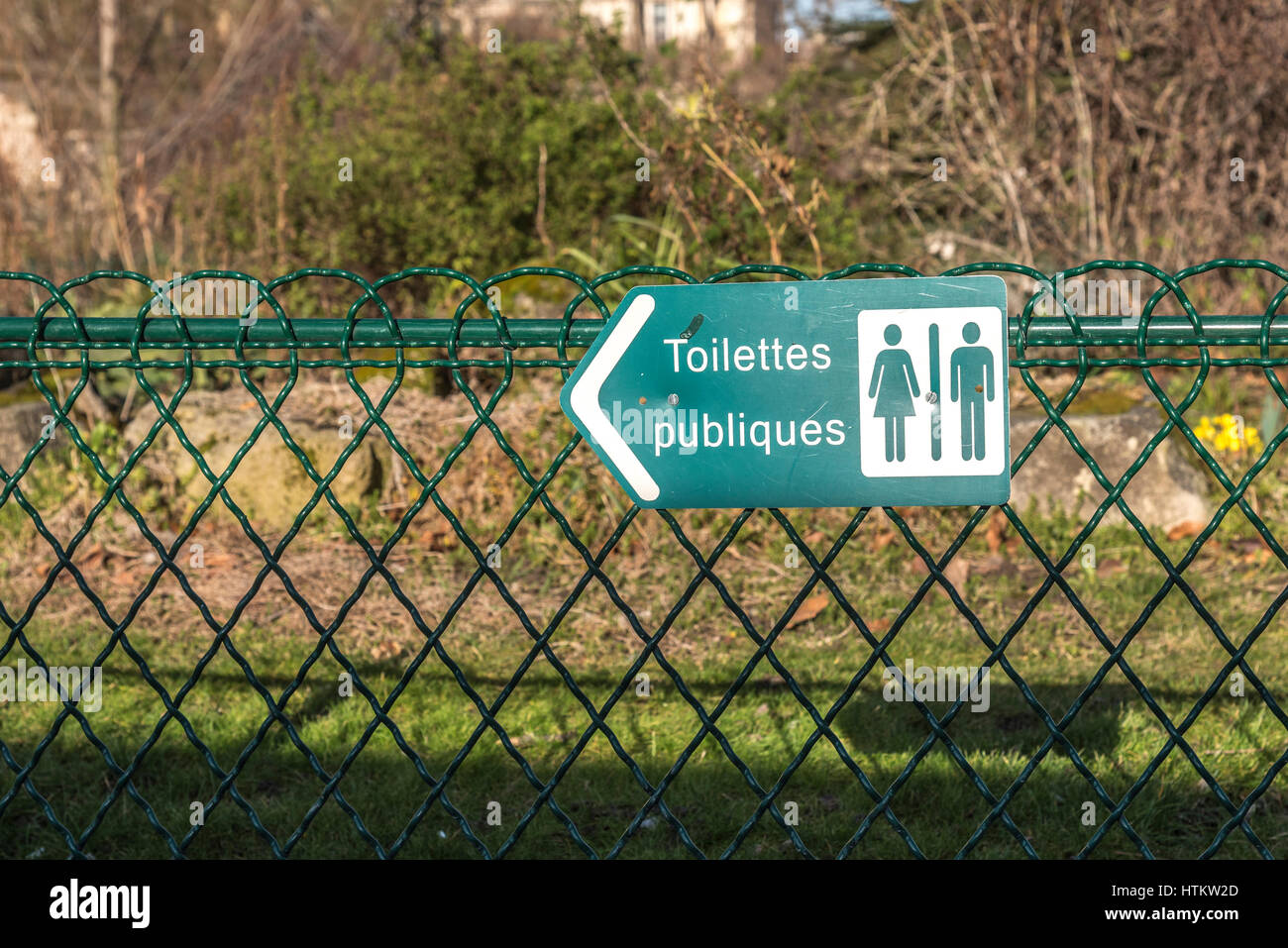 A French sign on a green wire fence points the direction to the mens ...