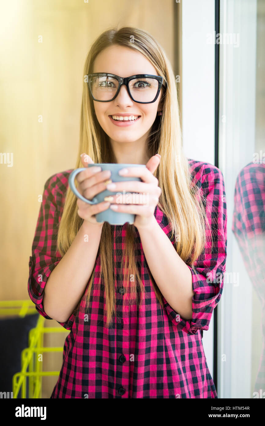 Young girl with cup of coffee with good mood in modern office near window Stock Photo