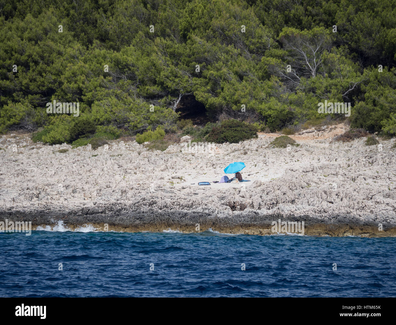 a lone single one sunbather alone on an isolated stony shingle rocky beach under the shade of a blue parasol umbrella in south Croatia Stock Photo