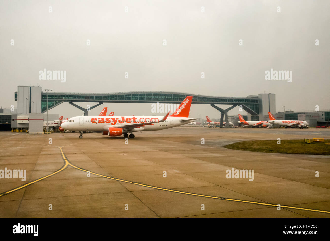 London Gatwick. England UK.  EasyJet aeroplanes on London Gatwick airport runway with a foot bridge that crosses the runway in the background.  There are airport buildings on both sides of the runway, as well as more aeroplanes parked at the terminal gates. Stock Photo