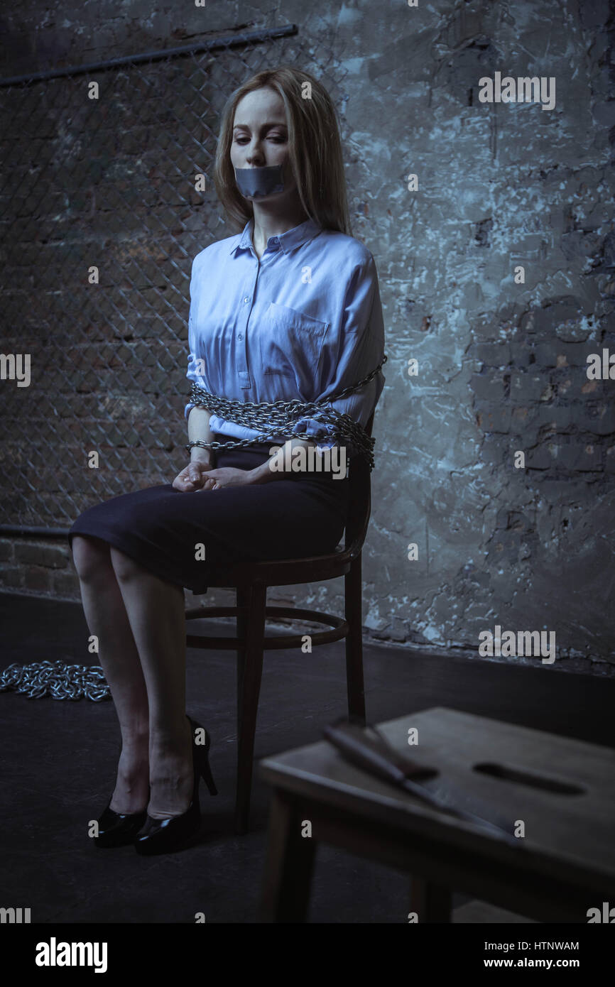 I wish I wasnt here. Anxious helpless bank worker sitting in a dark room being tied up to a chair while looking at the dagger lying nearby Stock Photo