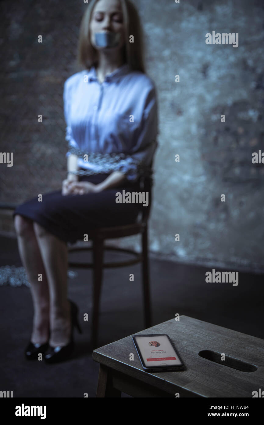 Important call. Trapped helpless miserable girl trying figuring out who calling while sitting in a dark room tied up to a chair Stock Photo