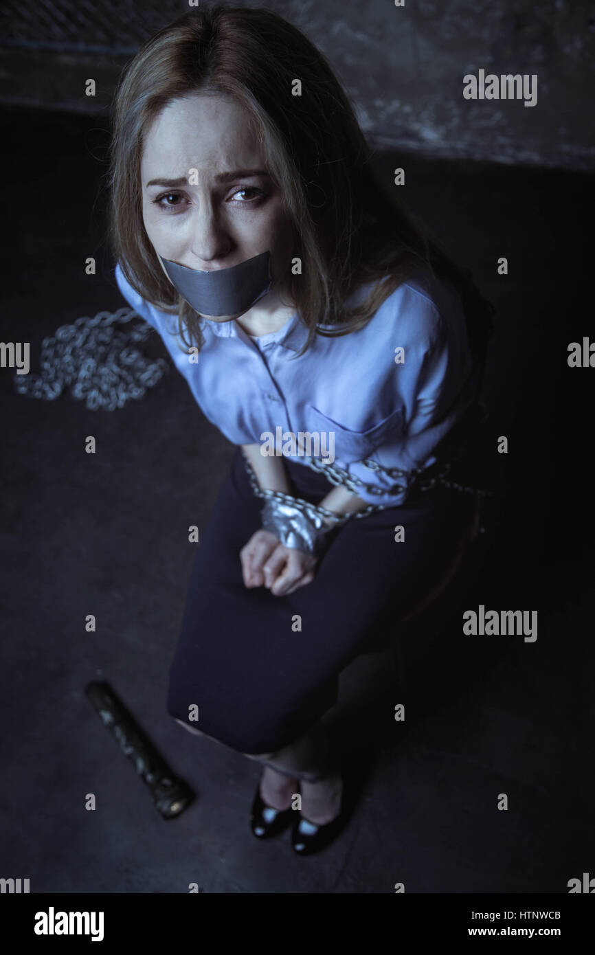 Help me, please. Trapped protectless weak girl looking desperate while sitting on a chair tied up with chains and her mouth shut with tape Stock Photo