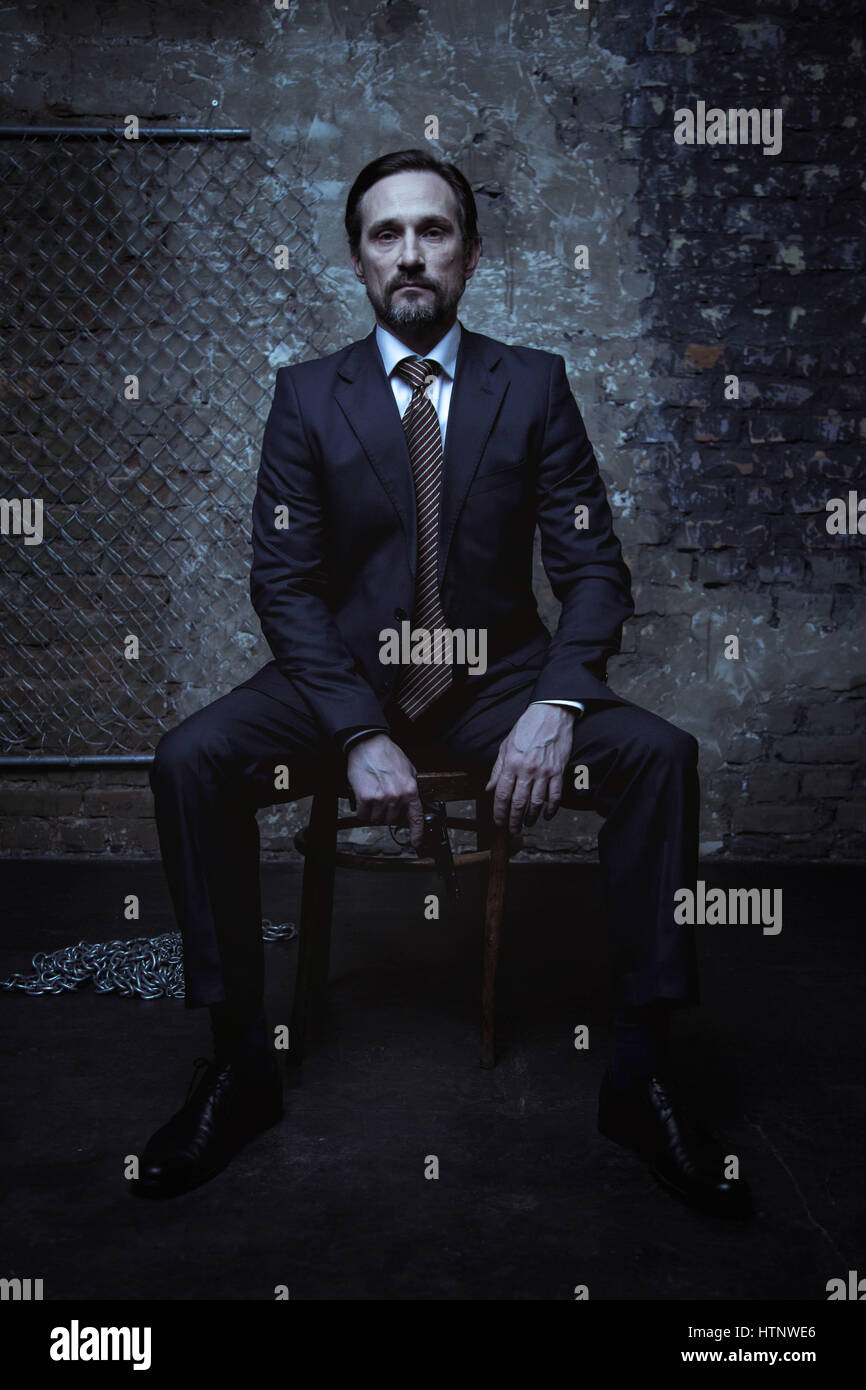 Super villain. Good looking brunette sharp gentleman wearing a classic suit and sitting on a chair in a dark room while holding a gun Stock Photo