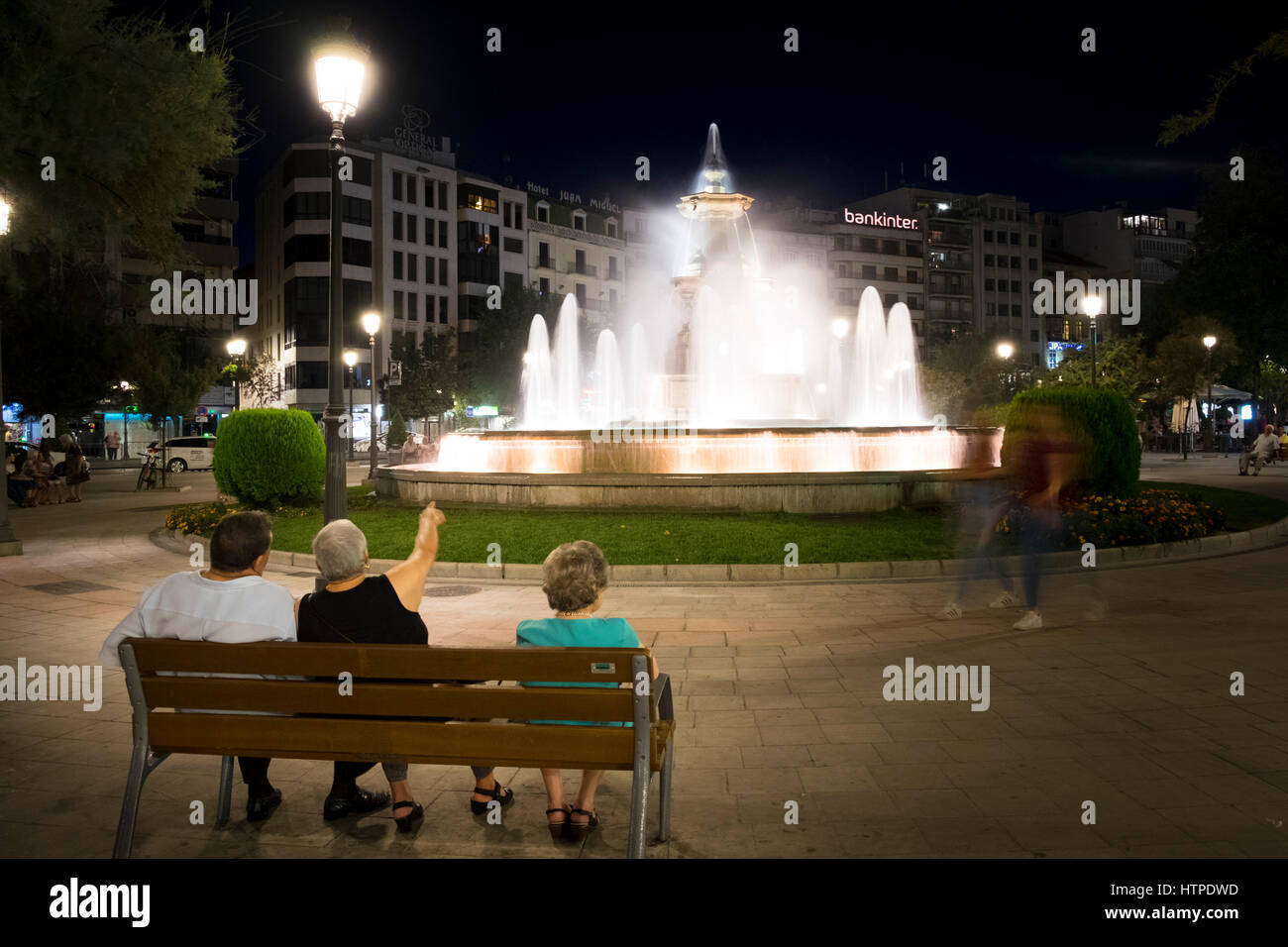 Three older people sit on a bench back to camera looking at the illuminated fountain in the Plaza del Campilo on a summer evening in Granada Spain Stock Photo