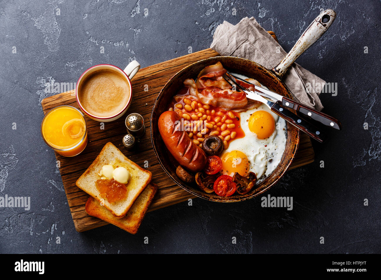 English Breakfast in cooking pan with fried eggs, sausages, bacon, beans, toasts and coffee on dark stone background Stock Photo