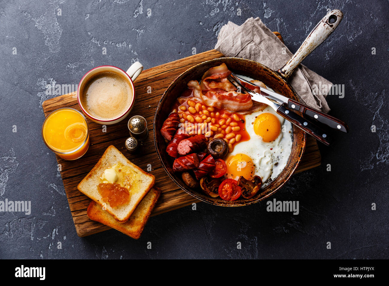 English Breakfast in cooking pan with fried eggs, sausages, bacon, beans, toasts and coffee on dark stone background Stock Photo