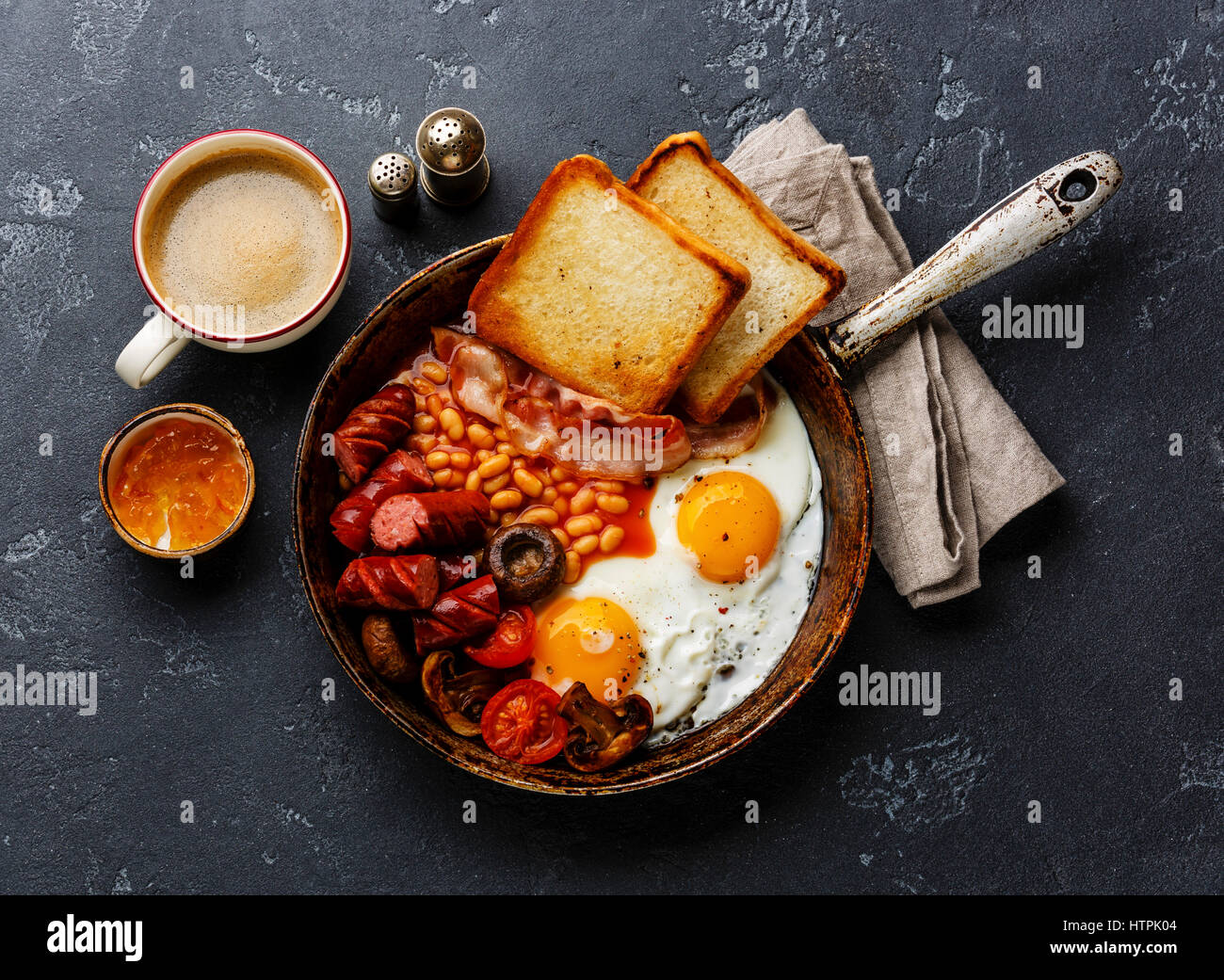 English Breakfast in cooking pan with fried eggs, sausages, bacon, beans, toasts and coffee on dark stone background Stock Photo