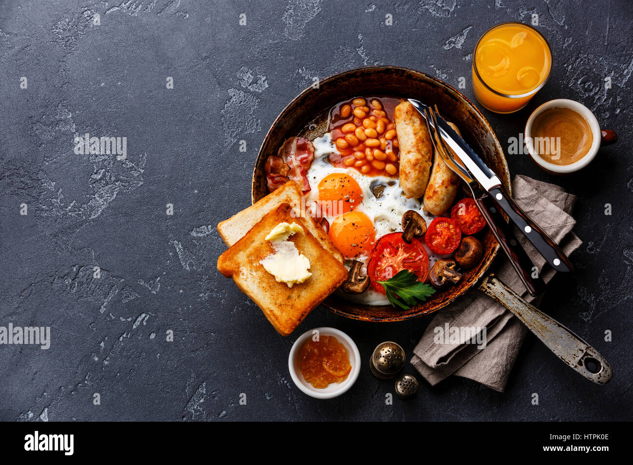 English Breakfast in cooking pan with fried eggs, sausages, bacon, beans, toasts and coffee on dark stone background copy space Stock Photo