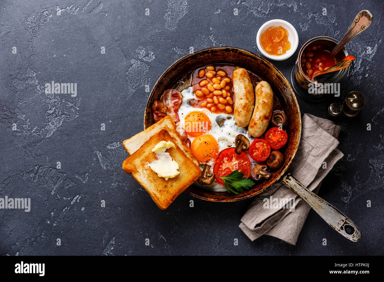 English Breakfast in cooking pan with fried eggs, sausages, bacon, beans and bread toasts on dark stone background copy space Stock Photo