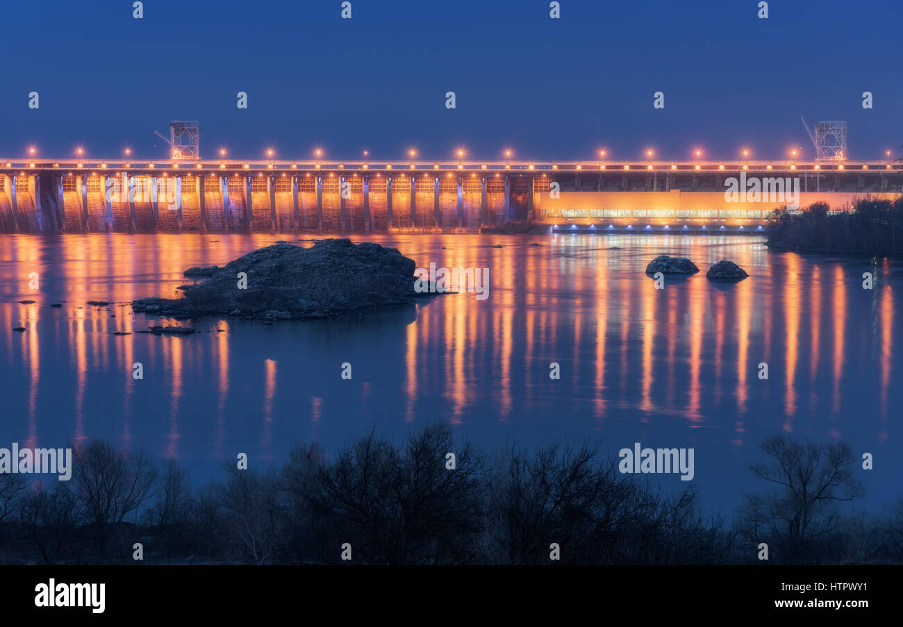 Dam at night. Beautiful industrial landscape with dam hydroelectric power station, bridge, river, city illumination reflected in water, rocks and sky. Stock Photo