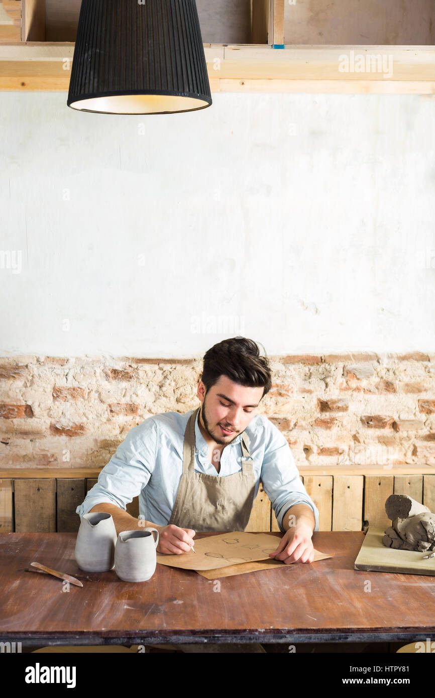 the smiling handsome young mexican potter does sketches of ceramics on the paper. concept of small business, handcrafted work. vertical picture with empty space for the text. Stock Photo