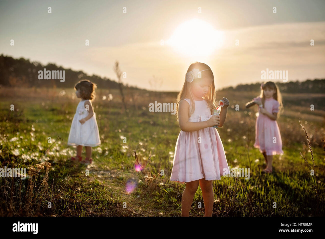 Three little girls in pink and white dress at the green field in sunset ...