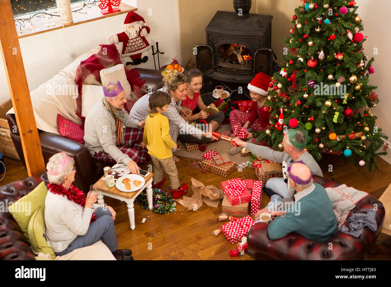 A family are sat around a christmas tree, opening presents and pulling crackers. Stock Photo